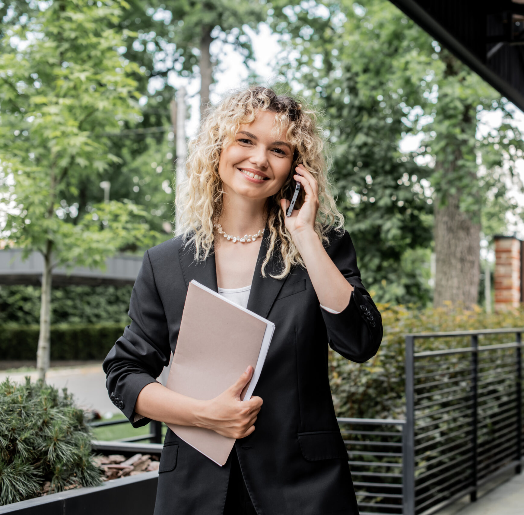 positive real estate agent with folder talking on smartphone and looking at camera near house